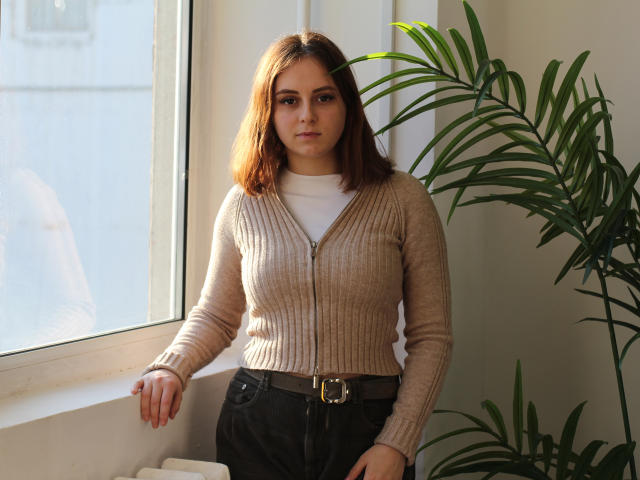 MilanaMiller has brown hair, a serious expression, and wears a beige ribbed cardigan with a zipper, black pants, and a white shirt. She's standing by a window with a plant.
