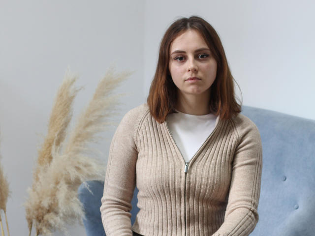 MilanaMiller with brown hair, wearing a beige ribbed cardigan over a white tee, sitting on a blue chair, looking serious, with puffy beige grasses to her left.