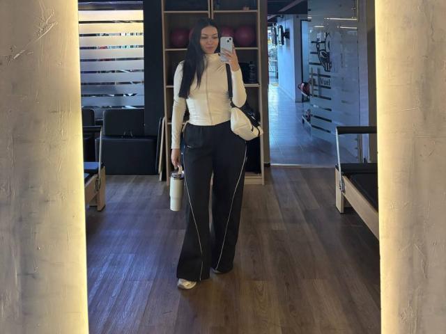 BerlyVenuss with long black hair, white blouse, black pants, holding phone in mirror selfie, standing in gym, wooden floor, white bag on right. Her ass is visible.