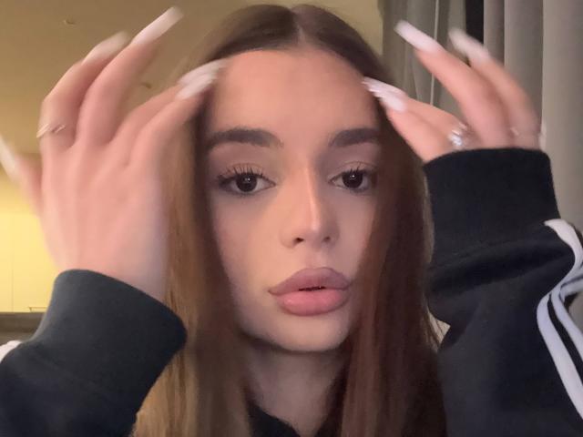 RoseleeVandiest with long brown hair, full lips, and big eyes, hands in her hair, wearing a black shirt with white stripes, has long, pointed nails, and a ring on her left hand.