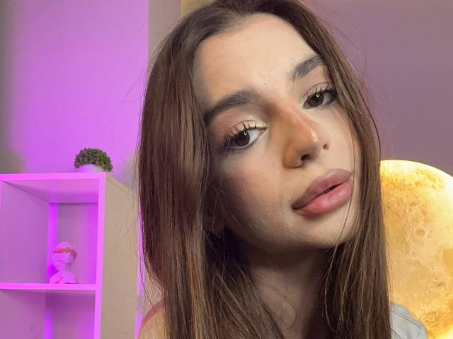 RoseleeVandiest with long brown hair, brown eyes, and pink lipstick, looking slightly to the side with a subtle smile. Her makeup includes gold eyeshadow and thick eyelashes.