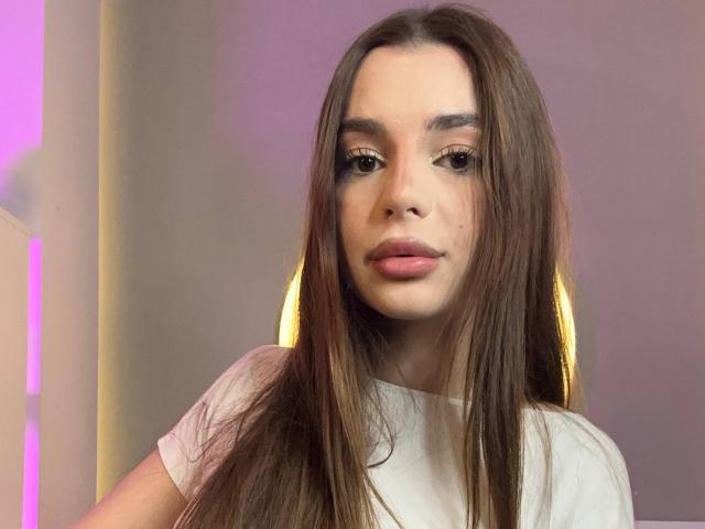 RoseleeVandiest has long brown hair, subtle makeup, and pinkish lips. She wears a white tee, sitting with a neutral expression. Her tits are small, and she's cute.