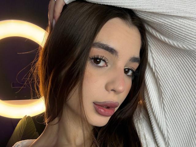 RoseleeVandiest with long brown hair, light skin, and full pink lips, holding a white ribbed shirt, has brown eyes with long lashes, and a subtle sexy smile.