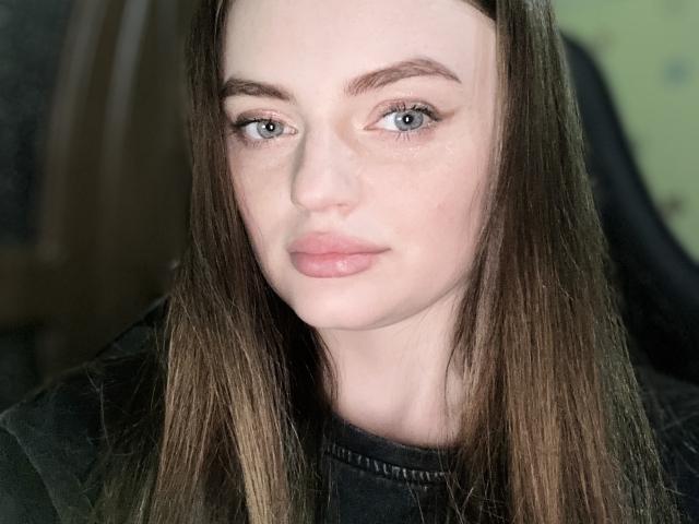 Anisa Healey tem pele clara, olhos azuis e cabelo castanho liso, veste uma camisa preta com ombros bufantes e tem lábios cheios e rosados, olhando diretamente para a câmera..
