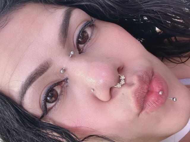 TarahDustin's face, with dark curly hair, brown eyes, and multiple facial piercings: nose, septum, and lower lip, showing a soft pink pout and slightly glossy lips.