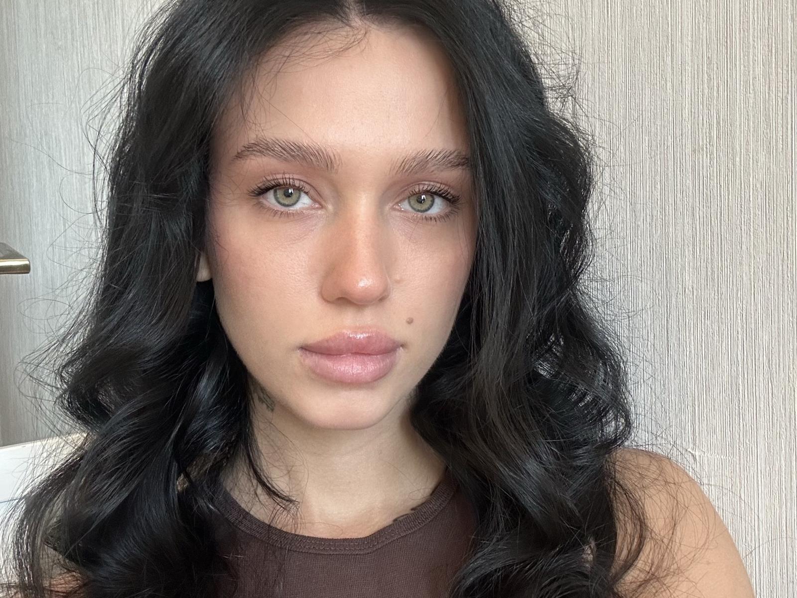 ShanikaBossick ha lunghi capelli neri ondulati, occhi verdi e labbra rosa lucide con una piccola lentiggine sulla guancia destra. Indossa una canotta marrone senza maniche..