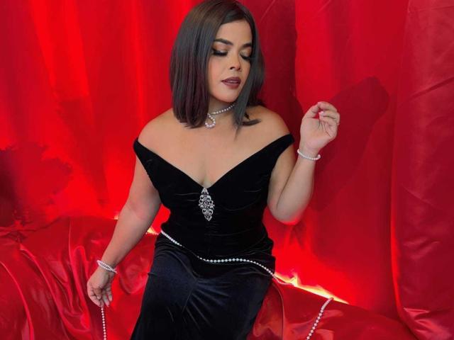 AbriilBeltran with medium tits, wearing an off-shoulder black velvet dress, silver necklace, bracelet, and pearl necklace, holding a pearl bracelet, against a red backdrop.