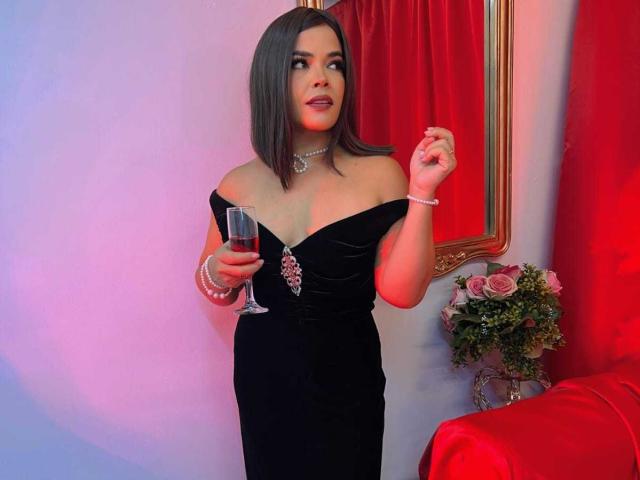AbriilBeltran with dark brown bob, black off-shoulder dress, pearl necklace, holding wine glass, red curtain, gold mirror, red chair, pink roses, and white wall in background.