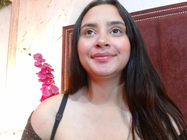 GraceBlue69 has long dark brown hair, brown eyes, and a cute smile. She's wearing a black off-shoulder top, showing her small tits. A pink orchid is in the background.
