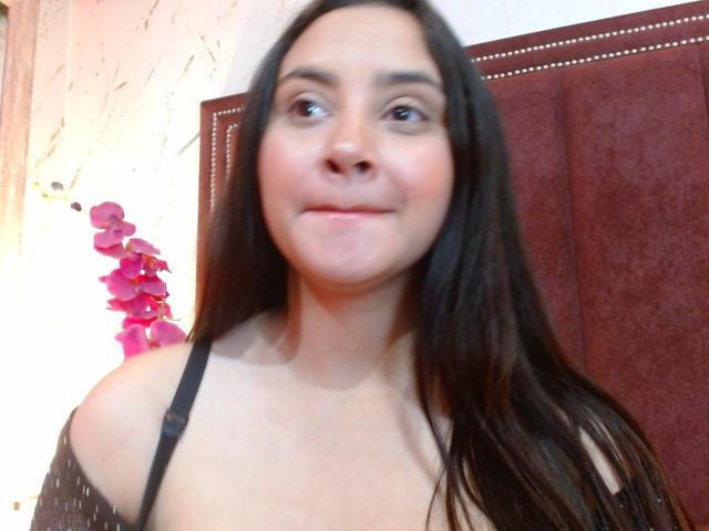 GraceBlue69 with long brown hair, light skin, and small tits, wearing a black strap, smiling slightly, and looking to the side, with a pink orchid in the background.