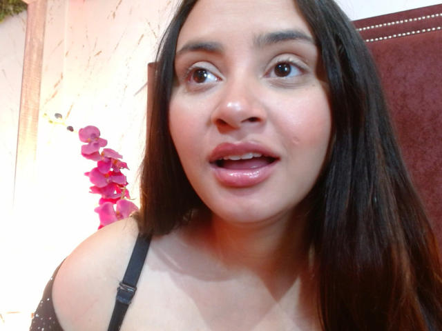 GraceBlue69 mit dunkelbraunen Haaren, leicht geöffneten, rosa Lippen, braunen Augen und einem schwarzen Trägerkleid, das ihre nackten Schultern zeigt und einen Hauch ihrer Brüste in einem Selfie..