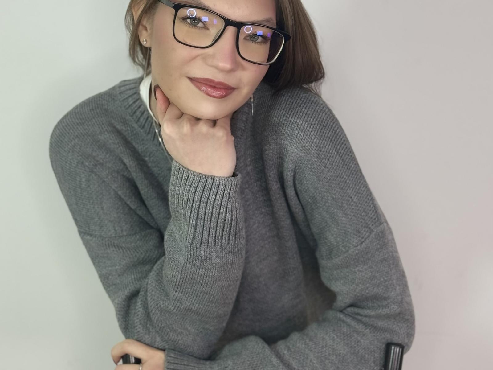 Jessica Harleston met een bril, grijze trui, hand op haar nek, bruin haar, rode lippenstift, een lichte glimlach, een witte muur, een zwarte polsband, zachte verlichting, kijkt omhoog, licht gekanteld hoofd..