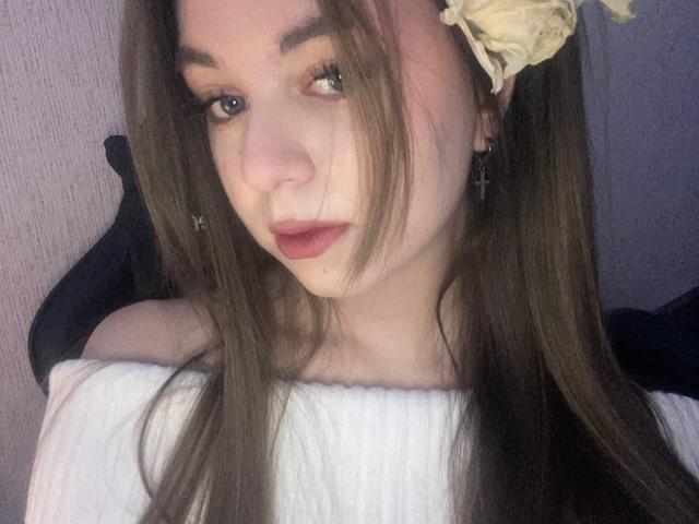 MartiSawdo with long brown hair, off-shoulder white top, red lipstick, cross earrings, and a yellow flower in her hair, looking slightly to the side, with soft lighting.