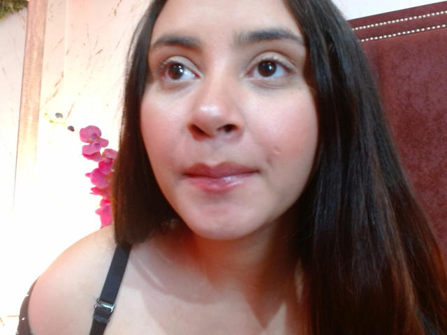 GraceBlue69 has long brown hair, brown eyes, and a slight smile with shiny lips. She's wearing a black strap, and her nose is slightly shiny.
