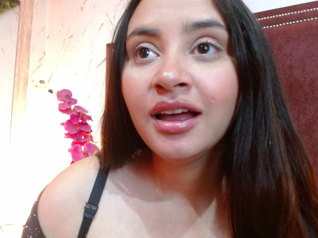 GraceBlue69 with medium brown skin, long black hair, brown eyes, slightly open mouth, wearing a black strap top, looking surprised. Pink orchid in background.