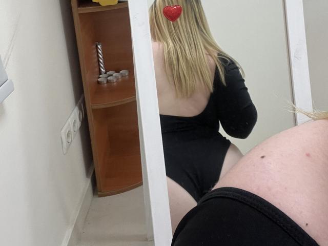 DiLaria with blonde hair, wearing a black, long-sleeve, open-back leotard, reflected in a mirror, showing her bare back and black thigh-highs, with a red heart sticker.