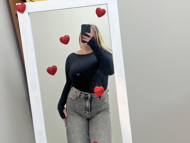 DiLaria's black long-sleeve top and gray high-waisted jeans, with red heart stickers covering her nipples and crotch, standing in front of a mirror, holding a phone.
