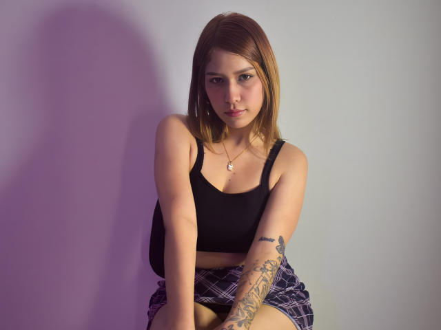 Sophie Saintclair, con cabello castaño claro, camiseta negra y falda escocesa, tiene un tatuaje en el brazo derecho y un collar, está sentada con una ligera sonrisa, mirando a la cámara..