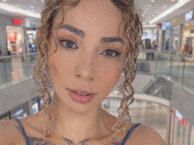 SilvanaXOwen has curly blonde hair, dark eyebrows, and wears a nose ring. Her blue eyes are slightly open, and she has a tattoo on her chest. She's in a mall.