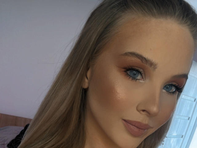 HeySiry has blue eyes, long blonde hair, and a cute face with subtle makeup, including brown eyeliner and mauve lipstick. She's looking at the camera with a slight smile.