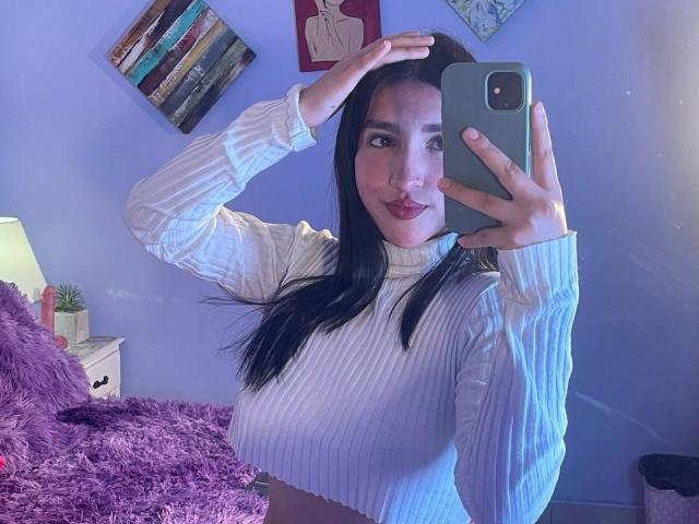 YviTaylor with long black hair, wearing a white ribbed sweater, taking a mirror selfie with her right hand on her head, showing her tits, and red lipstick.