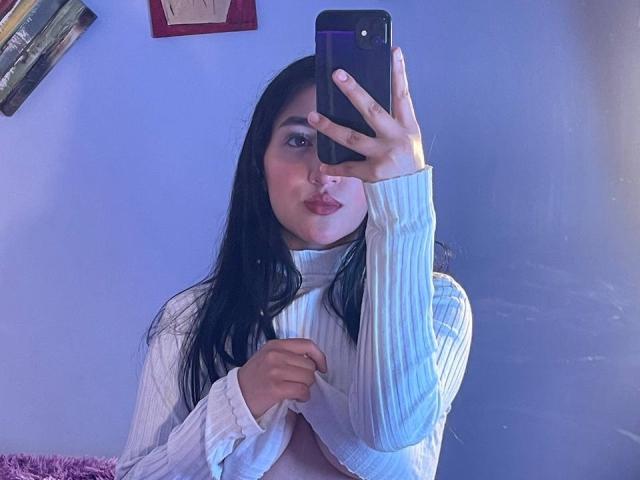 YviTaylor with long black hair, pouty lips, and white ribbed sweater, lifting shirt to reveal her tits, holding phone in mirror selfie.