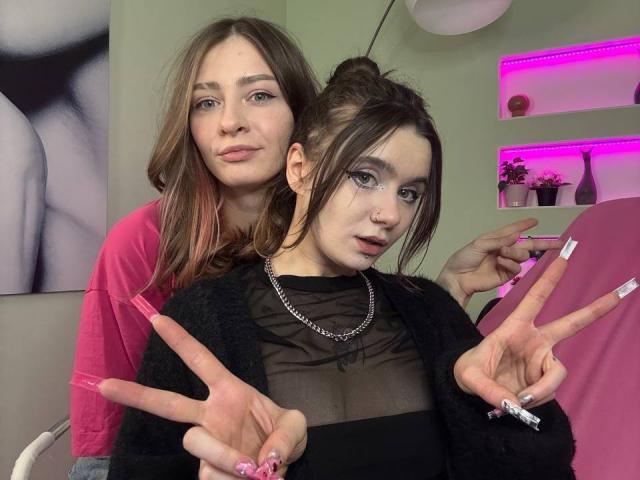LuluMao mit langen, braunen Haaren in einem pinken Oberteil steht hinter LuluMao mit einem schwarzen, transparenten Oberteil, pinken Nägeln und zwei Friedenszeichen. LuluMao im Vordergrund hat sichtbare Brüste und eine Nasenring..