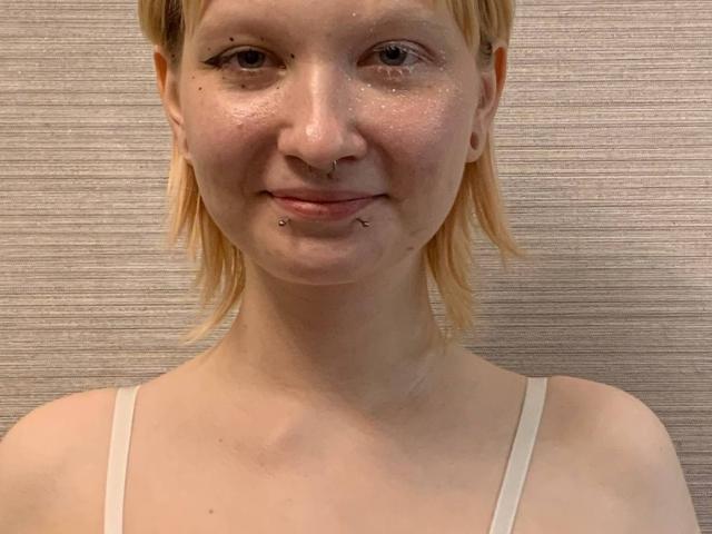 Jaditta, mit blonden Haaren, Nasenring und Piercing im Unterlippenbereich, trägt ein weißes Top mit Trägern und zeigt ein leichtes Lächeln mit subtiler Hautstruktur..