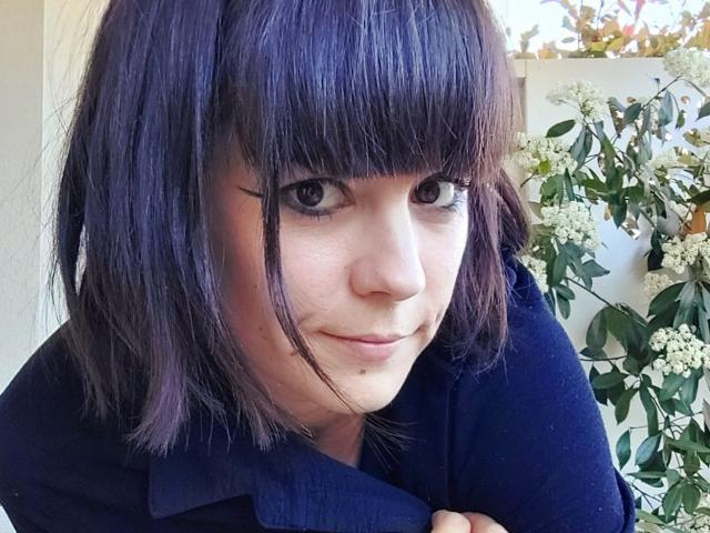OrianaLaFrancaise has dark purple, shoulder-length hair with bangs, black eyeliner, and is smiling slightly. She's wearing a dark blue shirt, looking directly at the camera.
