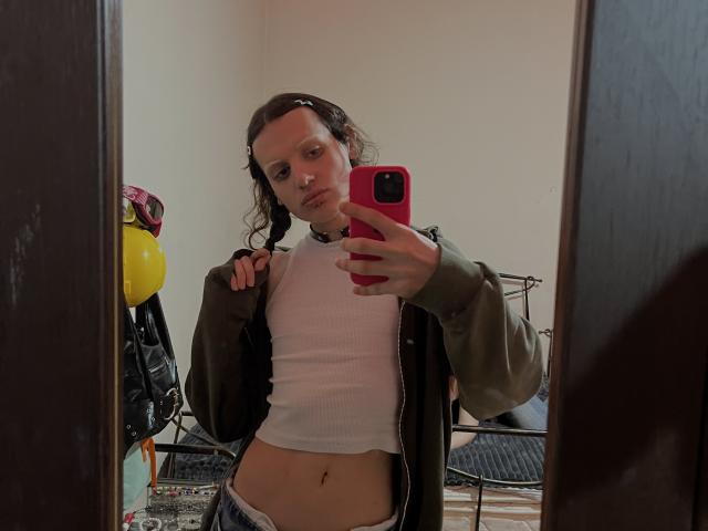 Sashenka69, avec des cheveux bruns bouclés, un débardeur blanc, une veste verte, tenant un téléphone rose, autoportrait devant un miroir, nombril visible, portant un jean bleu, le reflet montre une structure de lit en métal noir..