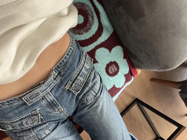De billen van Sashenka69 in blauwe spijkerbroeken, een wit shirt licht opgetrokken, waardoor een klein stukje van hun gebruinde huid zichtbaar is, zittend op een kleurrijke vloerkleed, naast een grijze stoel..