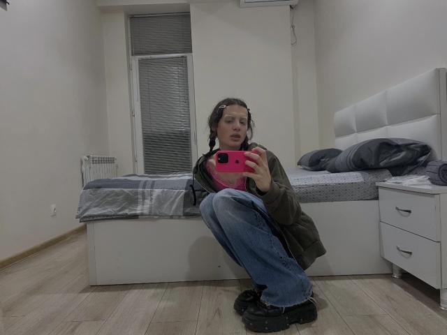 Sashenka69 zit in een hurk voor een spiegel, met een roze telefoon in haar hand, gekleed in een groene jas, blauwe jeans en zwarte laarzen, waarbij haar borsten en billen licht zichtbaar zijn..