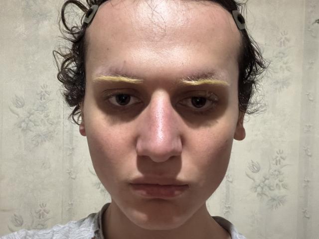 Sashenka69 tiene maquillaje amarillento en las cejas, cabello negro rizado con clips metálicos y una expresión neutra en un rostro pálido, ligeramente salpicado de pecas. La nariz de Sashenka69 es pequeña y ligeramente rosada..