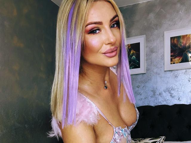 YaseminSaf with purple and blonde hair, pink eyeshadow, and a shiny nose, wearing a sparkly top, showing her tits, and a cute smile, in a modern room.