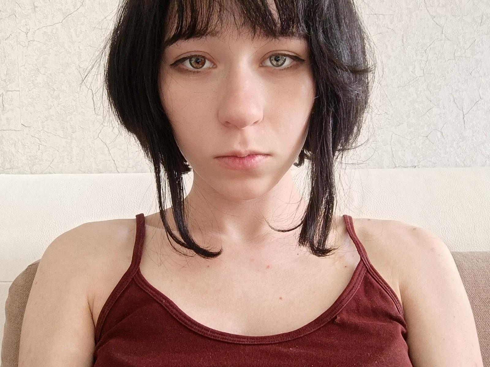 ChiSkocilich has short black hair, green eyes, and a serious expression. She's wearing a red tank top, showing her small tits and pale skin. She's sitting on a couch.