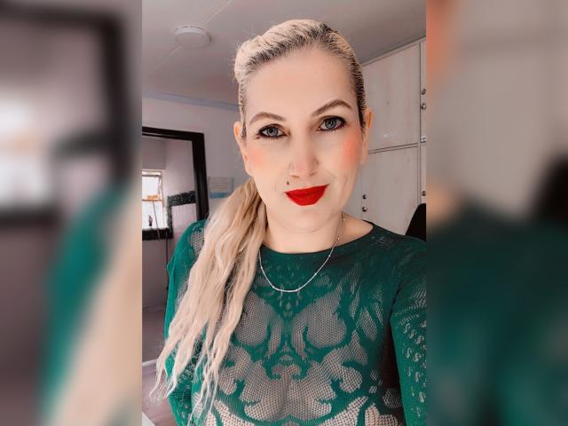  GiuliaAddams with blonde ponytail, green lace top, red lipstick, nose ring, and silver necklace, smiling at herself in mirror, holding phone. Her tits are visible through lace.
