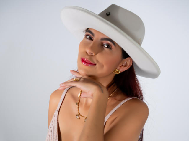 Carla Torres, con rossetto rosso, indossa un cappello bianco, orecchini dorati e una collana, mentre si tocca il collo, sorride, mostra il seno e indossa una maglietta rosa..