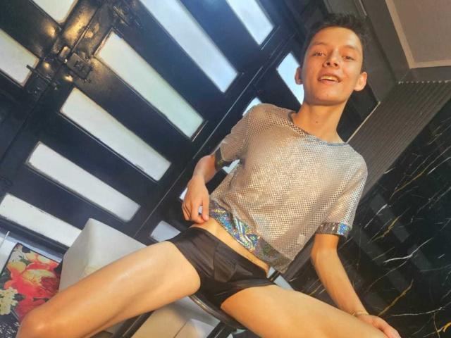 SamuJasper with short black hair, wearing a mesh gray shirt and black underwear, sitting with legs spread, smiling at the camera, showing a slim, cute, and sexy build.