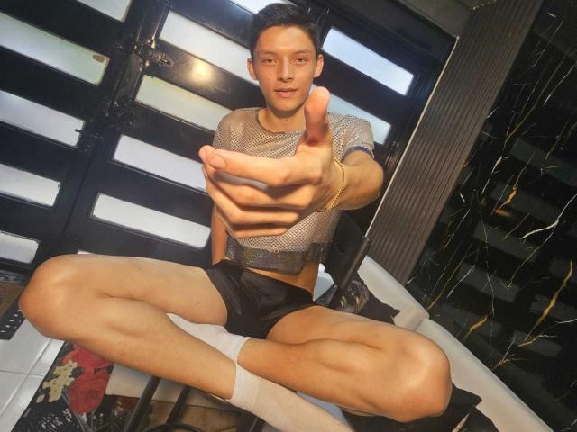 SamuJasper with a muscular build, wearing a mesh shirt, black shorts, and white socks, pointing forward with a serious expression, sitting with legs spread in a room.