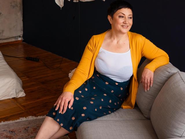 EmilyLlove with short black hair, yellow cardigan, white tank top, and blue skirt with peach patterns, sitting on gray sofa, smiling, showing her large tits, on wooden floor.