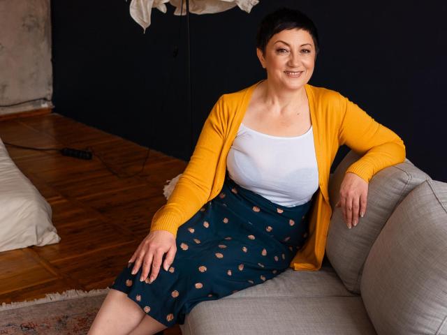 Emily, con senos grandes, vestida con un cárdigan amarillo, una camiseta blanca y una falda azul oscuro con lunares naranjas, sentada en un sofá gris, sonriendo..