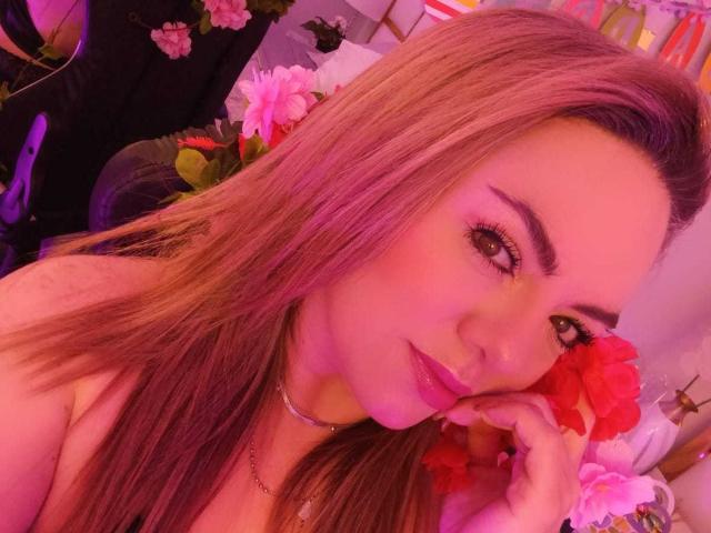 AvaMia with long, pinkish-brown hair, pink lipstick, and a flower in her mouth, holding her cheek, wearing a choker, in a pink-lit selfie. Her tits are barely visible.