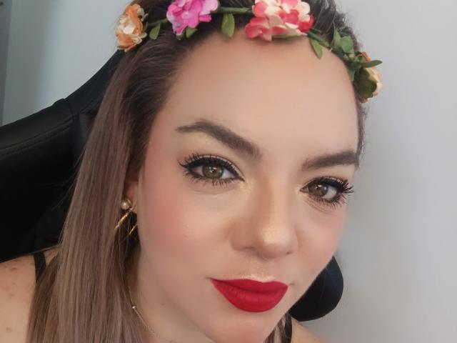 AvaMia with a flower crown, red lipstick, and thick black eyelashes, showing off her brown eyes and subtle makeup. She's got long brown hair and gold earrings.