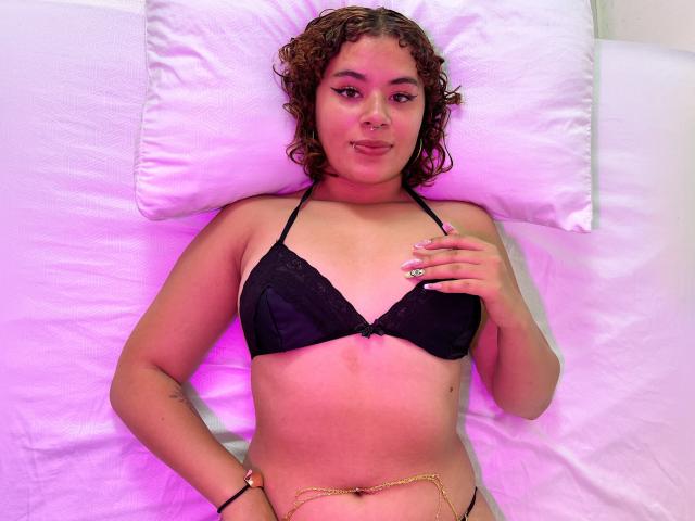 VeronicaMarie with curly brown hair, wearing a black lace bra, lying on a white pillow, has pink makeup, small gold chain, and a black thong, hand on chest.
