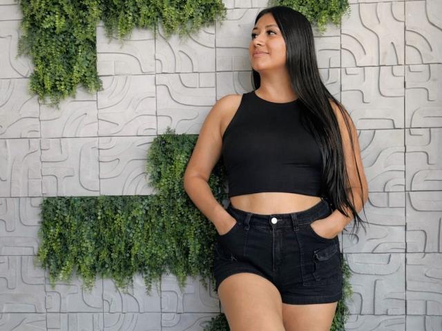 CatalinaRivero with long black hair, wearing a black crop top and high-waisted black shorts, hands in pockets, standing against a patterned wall with green hanging plants.