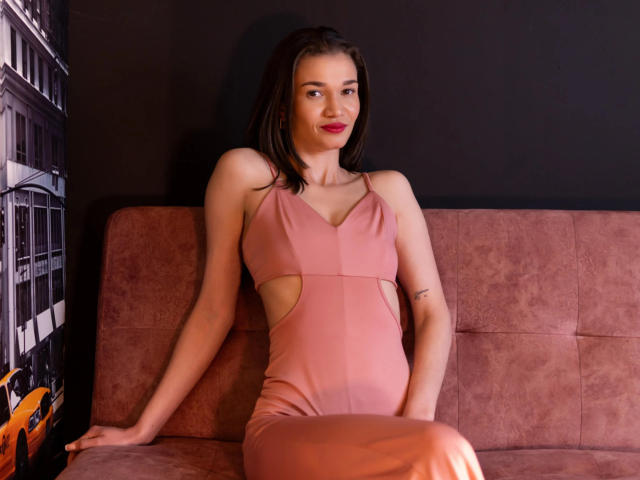 SammyTitss with black straps and nipple clamps on her big boobs, wearing a red collar and chain, has long black hair, and red lipstick, sitting on a purple couch.