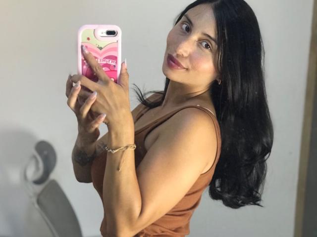 KateOwens with black hair, brown tank top, holding pink phone, smiling, showing her tits, in a mirror selfie, wearing a bracelet, pink nails, light skin, slight tattoo.
