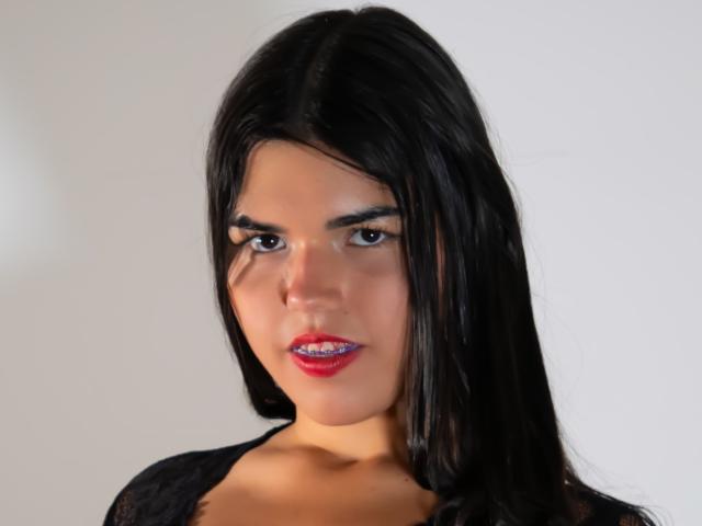 MiluJones has dark, straight hair, red lipstick, and is looking slightly to the side with a subtle smile. She has medium brown skin and is wearing a black lace top.