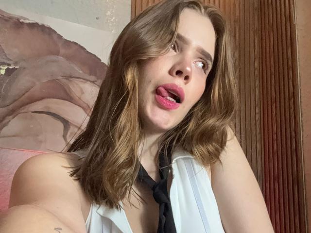 ArielLoveX with medium brown hair, pink lipstick, tongue out, white sleeveless top, black necktie, looking up, showing a tattoo on her right arm.
