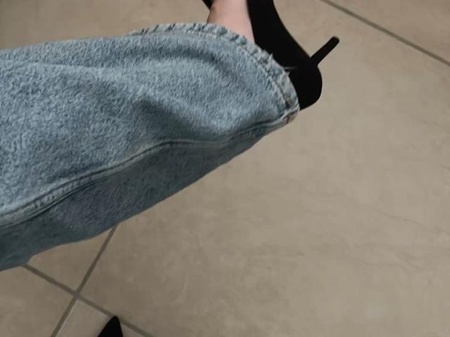 Lizee's denim-clad leg in black high heels, showing a slight bend, with her foot angled upward on a tiled floor.